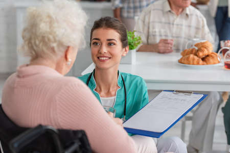 Smiling nurse holding clipboard near blurred senior woman in wheelchair in nursing homeの写真素材