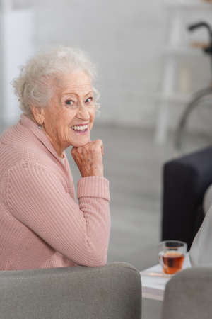 Elderly woman smiling at camera near blurred tea in nursing home