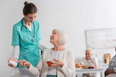 Smiling patient pointing at pills and holding tea near nurse in nursing home