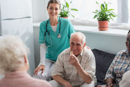 Smiling nurse sitting near senior interracial patients with chess figure in nursing home
