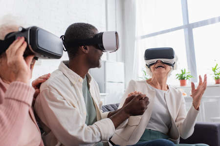 Cheerful senior woman holding hand of african american friend while gaming in vr headsets in nursing...