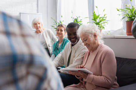 Smiling interracial pensioners using digital tablet near nurse in nursing home
