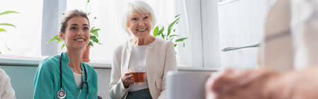 Smiling nurse and elderly woman with tea looking at man in nursery home, bannerの写真素材