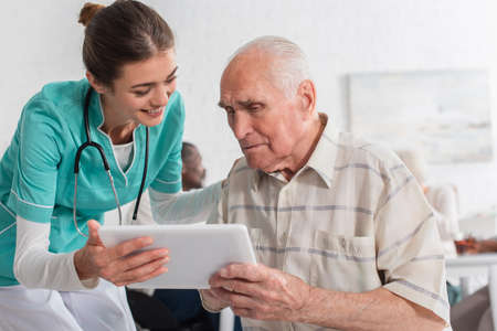 Positive nurse holding digital tablet near elderly man in nursing homeの写真素材
