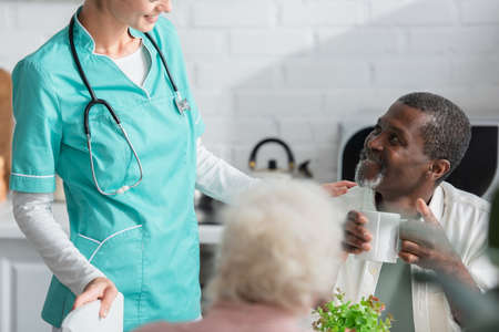 African american man holding cup near smiling nurse in nursing home