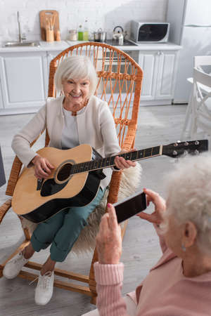 Smiling elderly woman playing acoustic guitar near friend with smartphone in nursing homeの写真素材