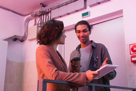 smiling african american technician with paper cup pointing at digital tablet while working with...