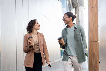 cheerful multiethnic friends with paper cups talking while walking on urban streetの写真素材
