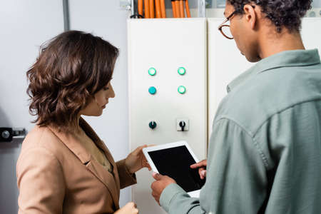 african american engineer pointing at digital tablet near colleague and switchboard of cooling...
