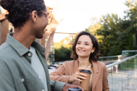 young woman with coffee to go smiling near blurred african american friend outdoorsの写真素材