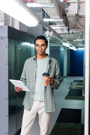 positive african american administrator with digital tablet and coffee to go looking at camera in...