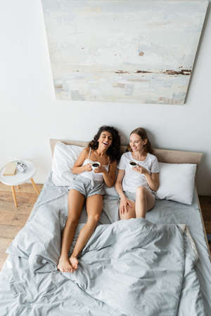 high angle view of happy lesbian couple holding cups of coffee in bed