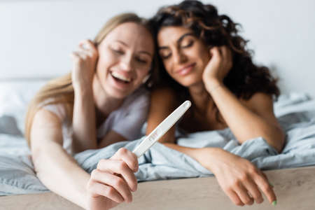 blurred and cheerful woman holding pregnancy test near curly girlfriend in bedroomの写真素材