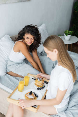 pleased young woman holding tray with tasty breakfast near curly girlfriend in bedの写真素材