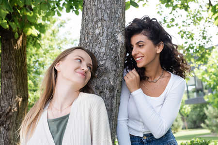 happy lesbian women looking at each other near tree trunk outside