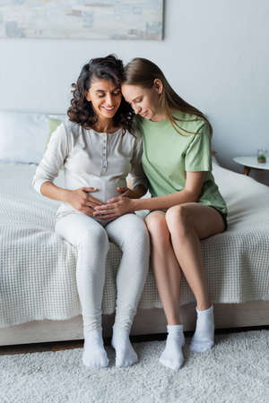 cheerful woman touching belly of pregnant girlfriend in bedroomの写真素材