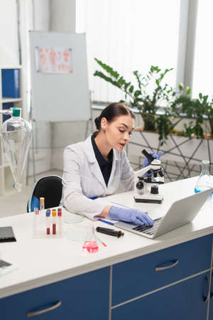 Brunette scientist using laptop near microscope and equipment in labの写真素材