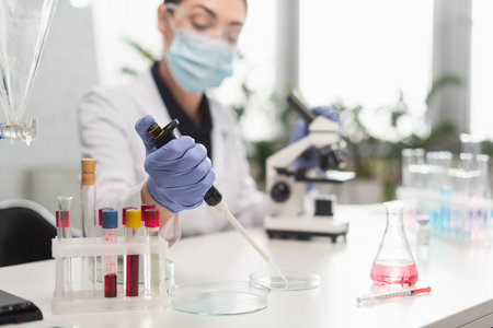 Blurred scientist in latex glove working with electronic pipette and petri dishes near test tubes...