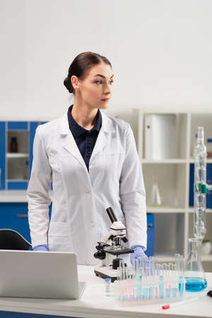 Scientist in white coat standing near medical equipment, laptop and vaccine in laboratory