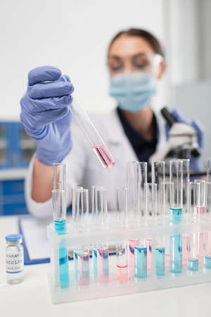 Blurred scientist in latex gloves holding test tube near coronavirus vaccine in lab