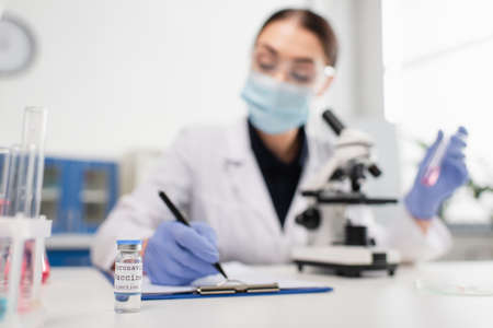 Coronavirus vaccine near blurred scientist writing on clipboard near microscope in labの写真素材