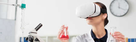 Scientist in vr headset holding flasks near microscope and equipment in laboratory, bannerの写真素材