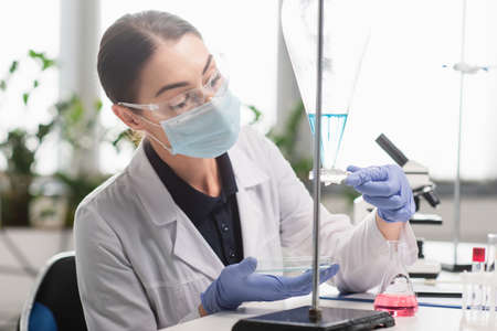 Brunette scientist in goggles and medical mask holding petri dish near flasks in lab