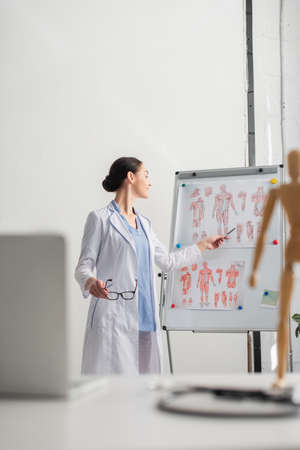 Side view of smiling doctor with eyeglasses pointing at flip chart near blurred laptop on table in...