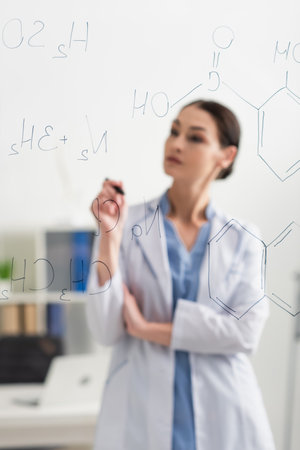 blurred doctor in white coat standing near chemical formulas on glass board