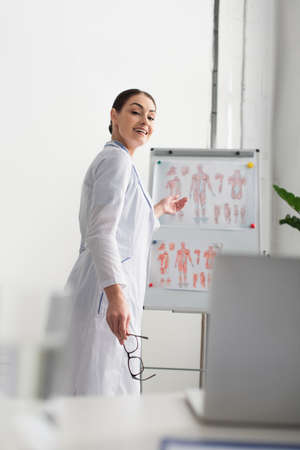 Smiling doctor holding eyeglasses and pointing at flip chart during video call on blurred laptop in hospitalの写真素材