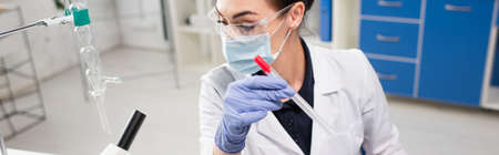 Scientist in medical mask holding pcr test near microscope in lab, banner