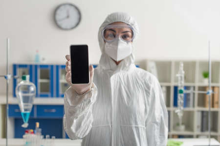blurred doctor in hazmat suit holding mobile phone with blank screen in labの写真素材