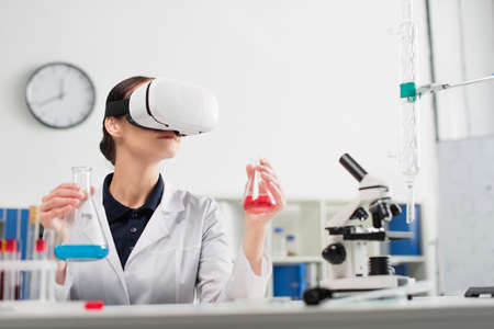 Scientist in vr headset holding flasks while working near microscope and test tubes in laboratoryの写真素材