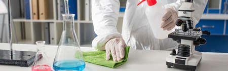 cropped view of doctor in hazmat suit wiping desk near flasks and microscope, banner