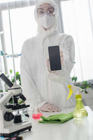 blurred scientist in personal protective equipment showing smartphone with blank screen near...
