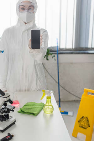 scientist in hazmat suit and goggles showing smartphone with blank screen in labの写真素材