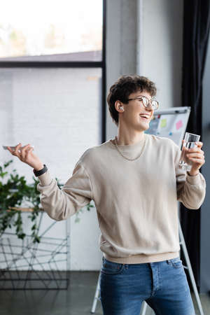 Smiling businessman in earphone holding smartphone and glass of water in office