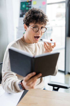 Astonished businessman recording voice message on smartphone and looking at blurred notebook in officeの写真素材