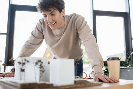 Cheerful businessman looking at model of building near eyeglasses and coffee to go in office