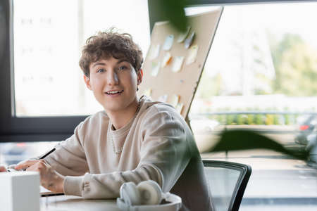 Positive transgender person looking at camera near blurred headphones in office