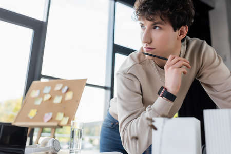Pensive businessman holding pen near blurred model of building and headphones on table in officeの写真素材
