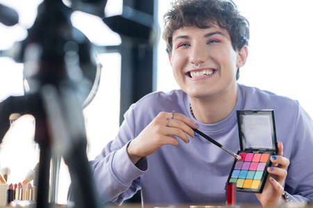 Smiling transgender person holding eye shadows and cosmetic brush near tripod and mirror