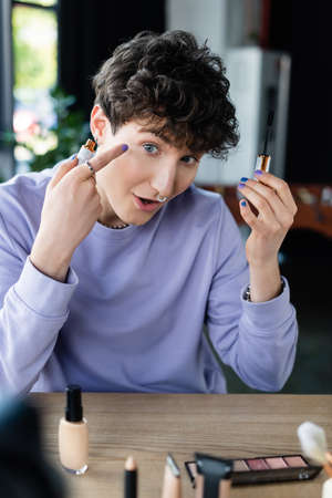 Transgender person holding mascara and pointing at eye near decorative cosmetics