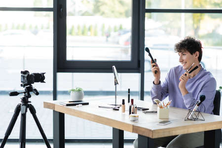 Cheerful transgender makeup artist holding cosmetic brushes near cosmetics and digital cameraの写真素材