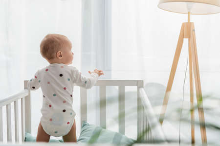 back view of toddler kid in baby romper standing in crib