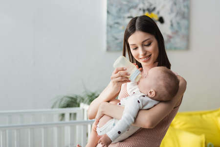 cheerful woman holding baby boy and feeding him with milk at homeの写真素材