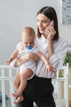 smiling woman talking on smartphone while holding baby boy in handsの写真素材