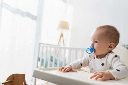toddler child with pacifier sitting in baby chair near blurred cribの写真素材