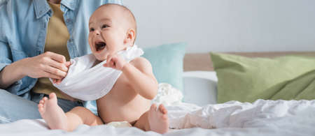 excited toddler with open mouth near mother dressing him on bed, banner