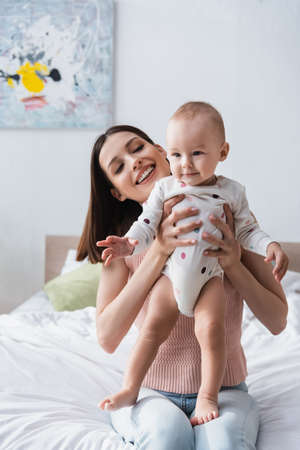 happy brunette woman holding toddler boy in romper while sitting on bed